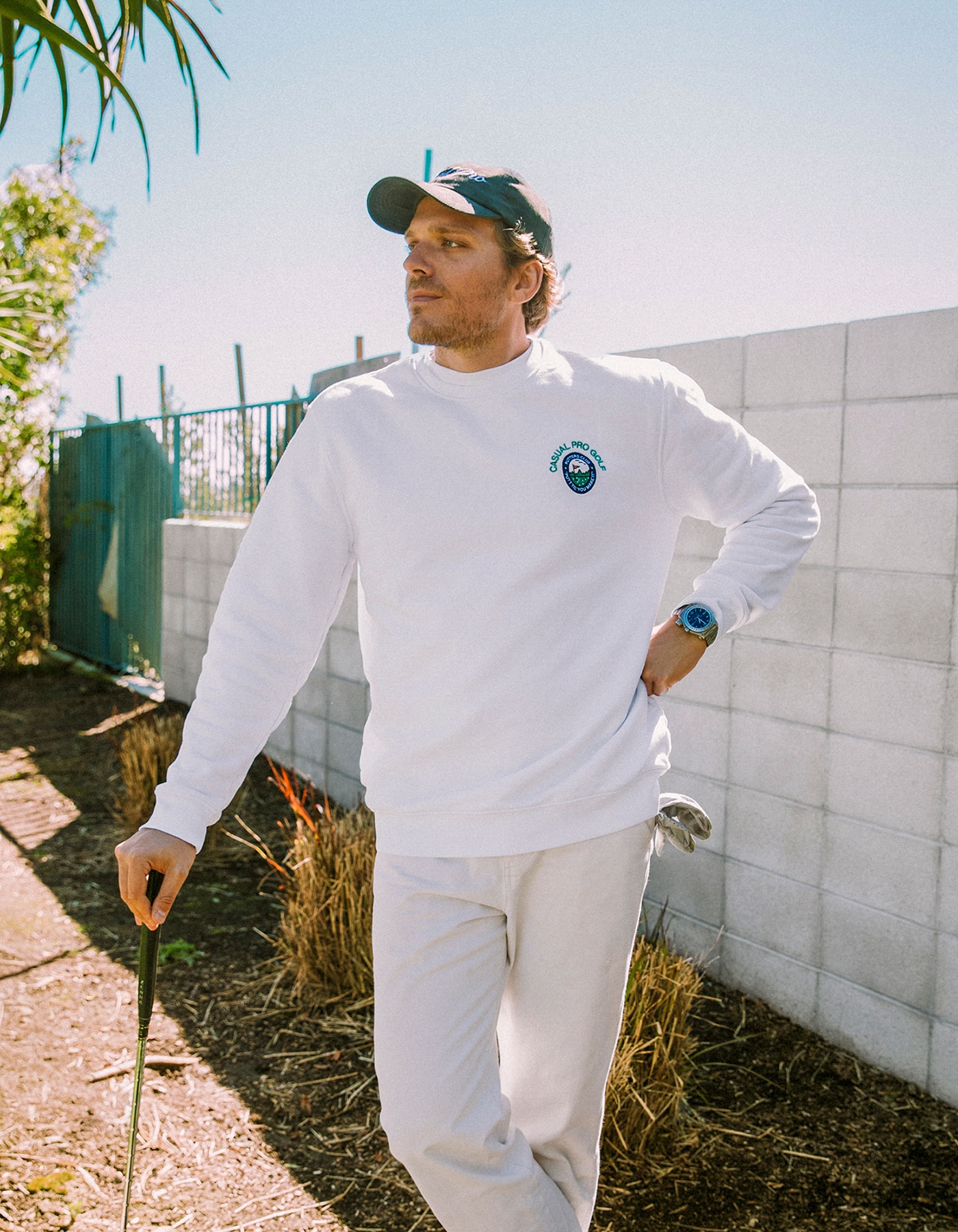 Man in white Casual Pro sweatshirt holding a golf club beside a concrete wall